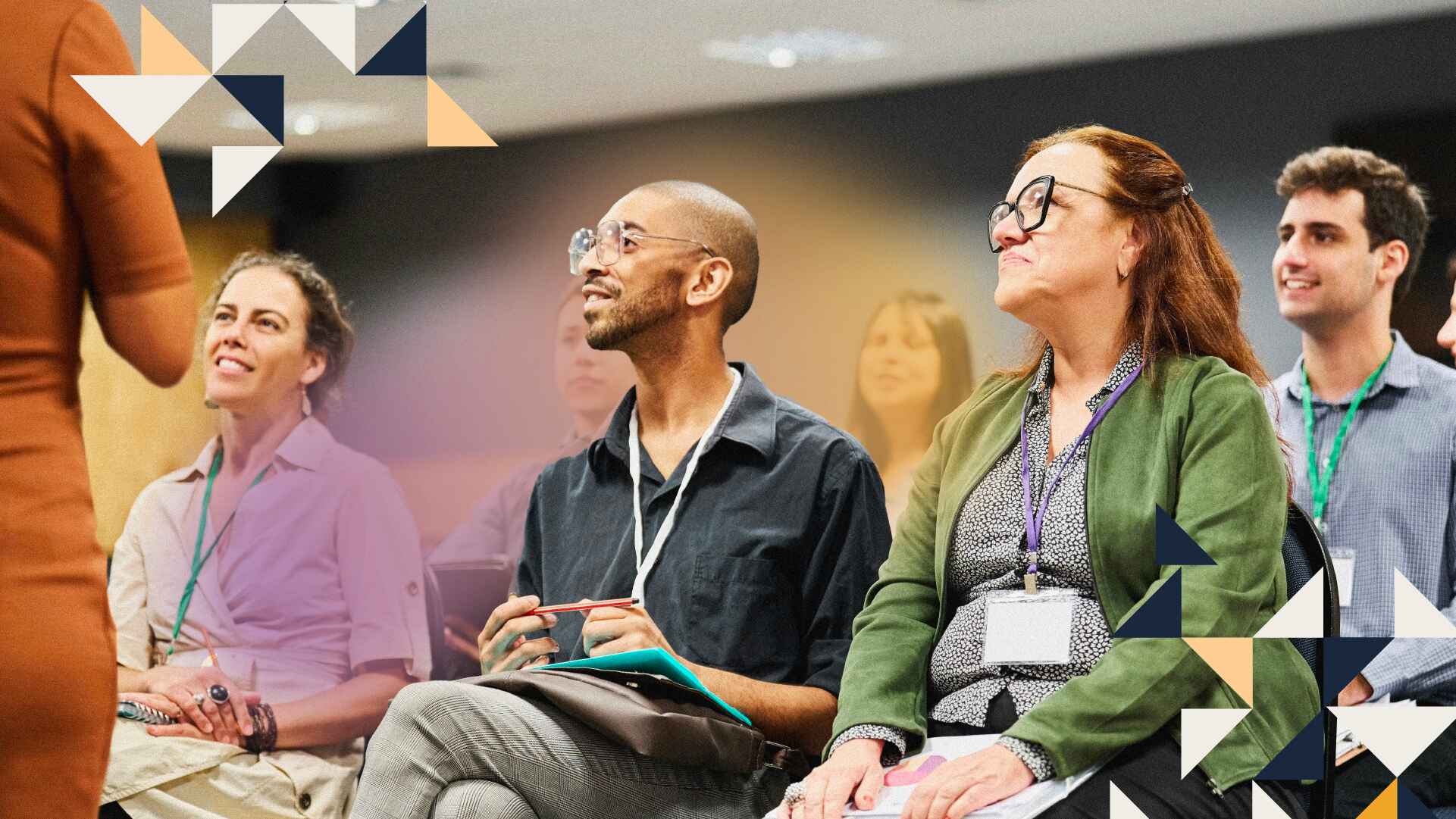 Two people sitting in an audience listening to a speech with event tags around their neck.